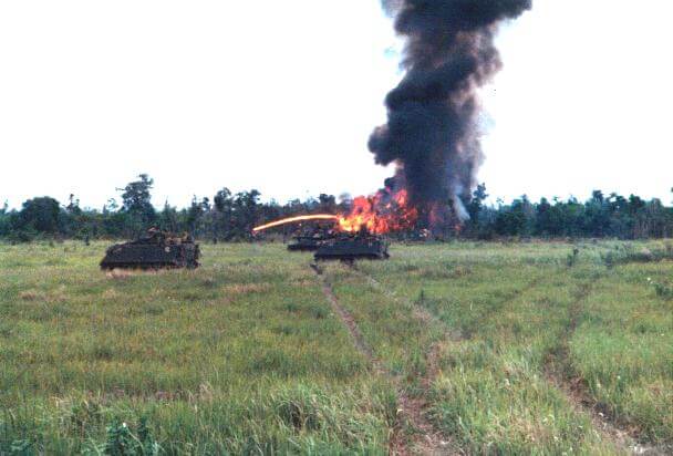 Tanks in a field, fire and smoke rising up from the jungle.
