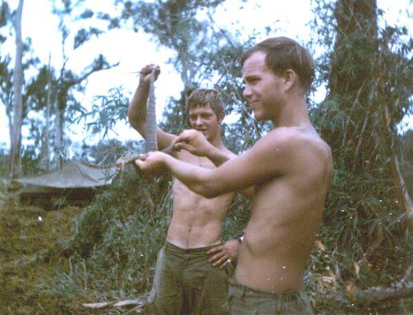 Two young shirtless soldiers in the jungle, holding up something for inspection.