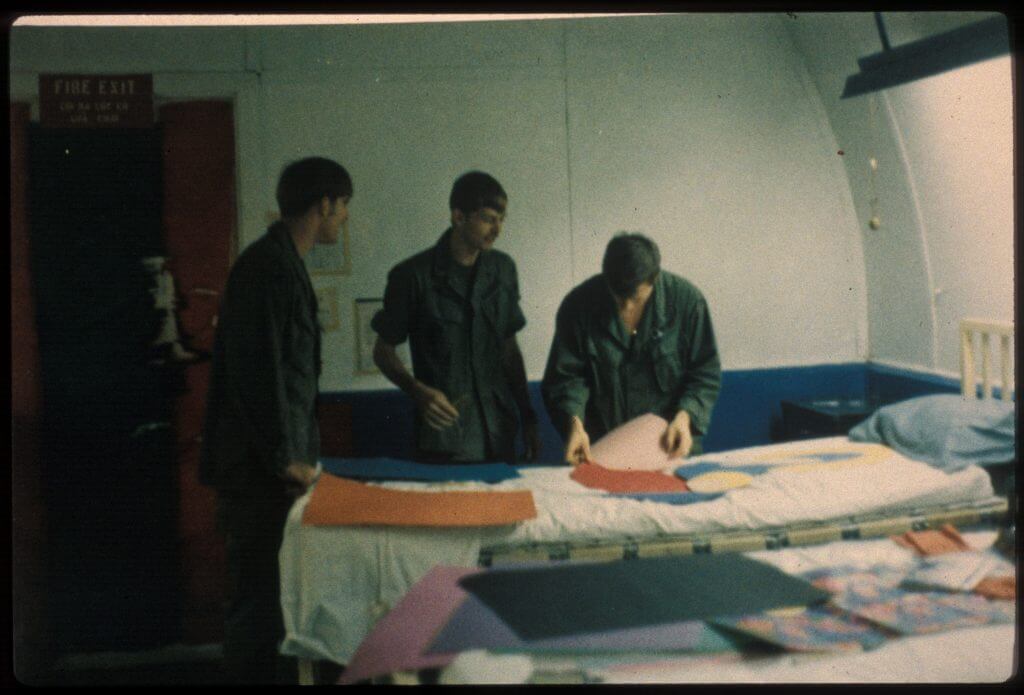 Three young men cutting construction paper, using a hospital bed as a work surface.