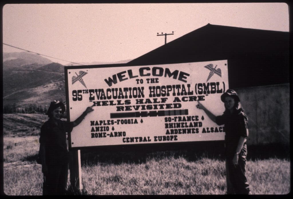 Two female nurses pointing to the welcome sign, which reads "Welcome to the 95th Evacuation Hospital - SMBL - "Hells Half Acre Revisited"
