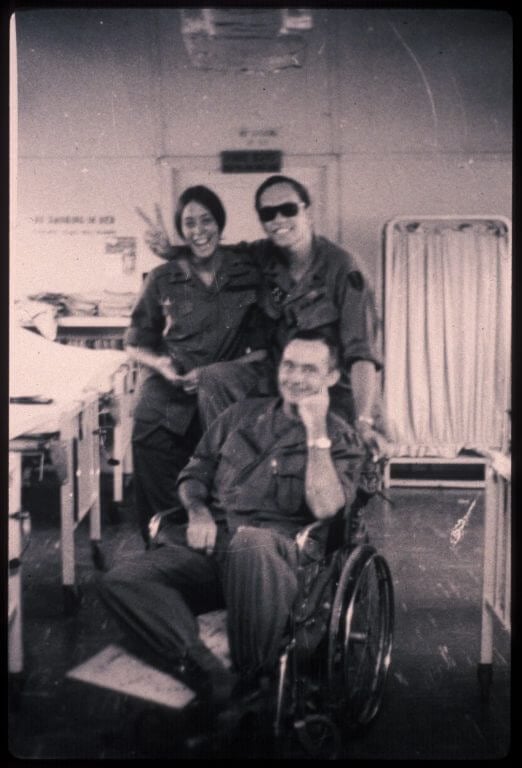 A smiling trio: a caucasian man in a wheel chair with a smiling caucasian nurse and an asian man in sunglasses putting up a peace sign.