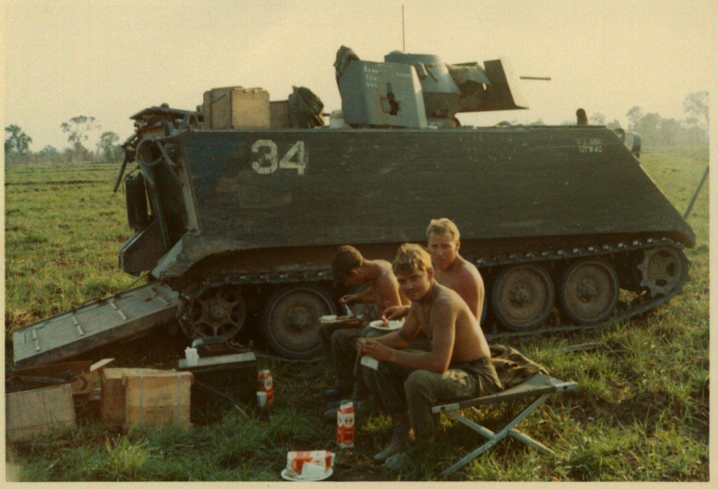 Three soldiers eating alongside a tank, in a field.