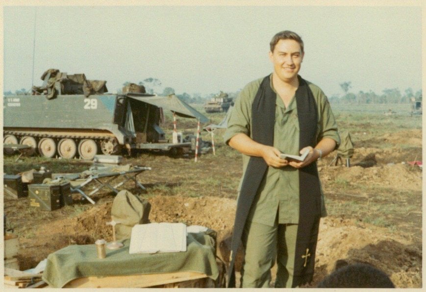 A young chaplain holding a small book, standing next to a makeshift alter in a field; tanks in the background.