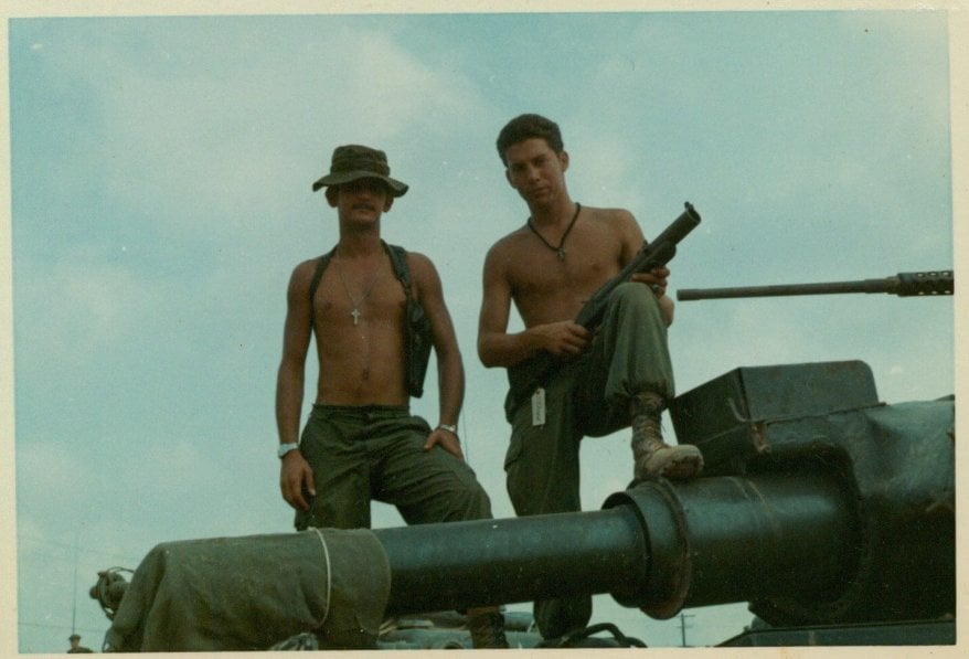 Two shirtless soldiers standing atop a tank near the guns, one holding a firearm.
