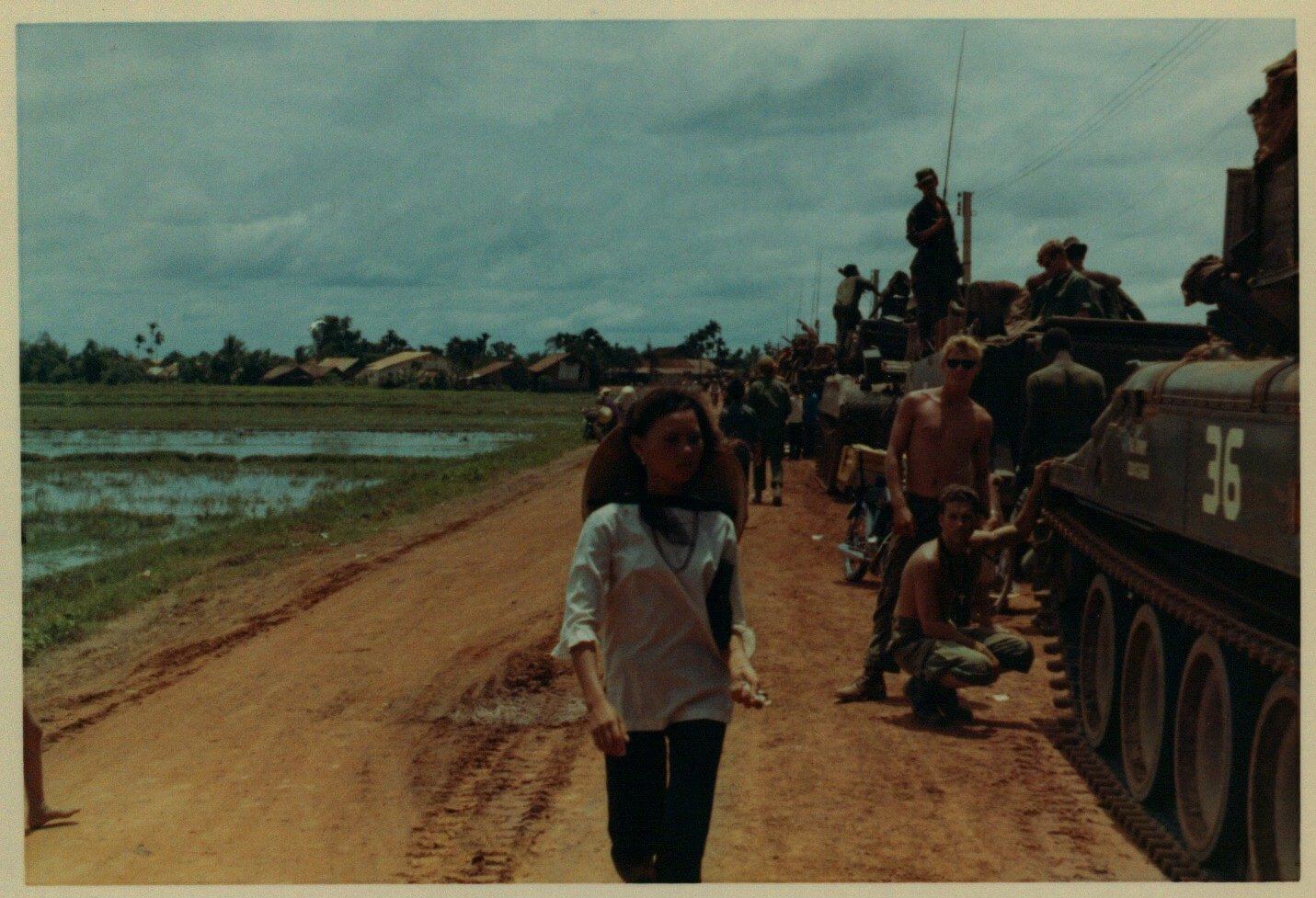 U.S. soldiers stand and lean on tanks watching a young Vietnamese woman walk by.