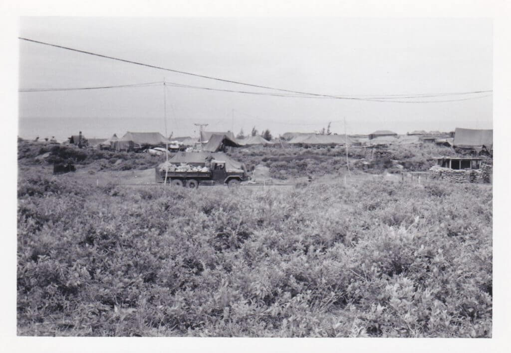 Landscape photo, brush in the foreground, a truck in the middle ground, and rooflines in the background.
