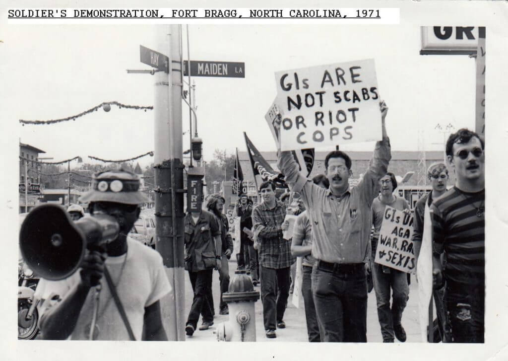 Male soldiers, black and white, protesting in the street holding signs and a megaphone.