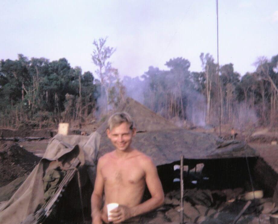 Young, shirtless U.S. soldier stands outside, smiling and holding a cup.