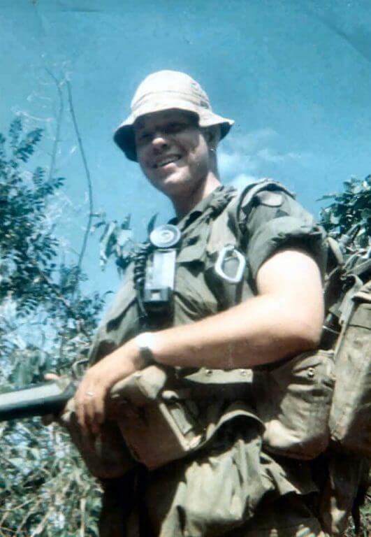 Young soldier in boonie hat, smiling, with radio hanging at his waist.