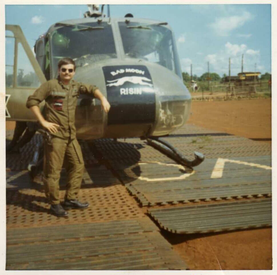 Young soldier in uniform and aviation sunglasses leaning against a helicopter with the text "Bad Moon Risin" on its nose.