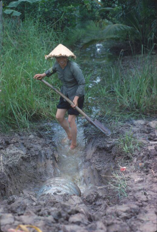 Woman with straw hat standing in a muddy ditch with a hoe.