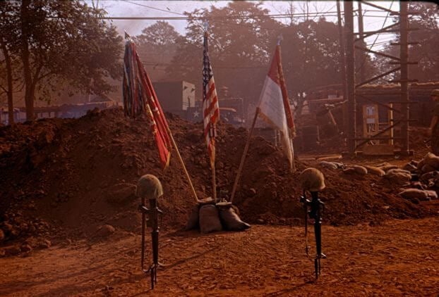 Rifles planted in the ground with helmets on top, flags in the background.