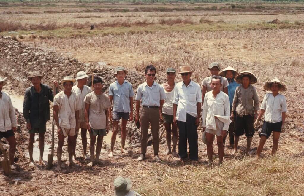 Group of about a dozen Asian men, barefoot, taking a break from working in the mud to pose for a picture.