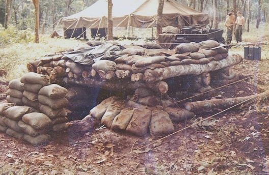 Bunker made of sandbags and logs. A group of shirtless soldiers stand far off in the background.