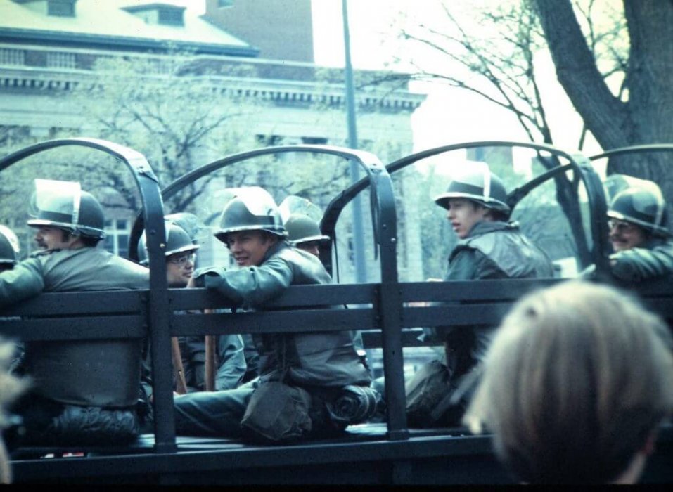 Guardsmen in riot gear sitting on the bed of a military vehicle.