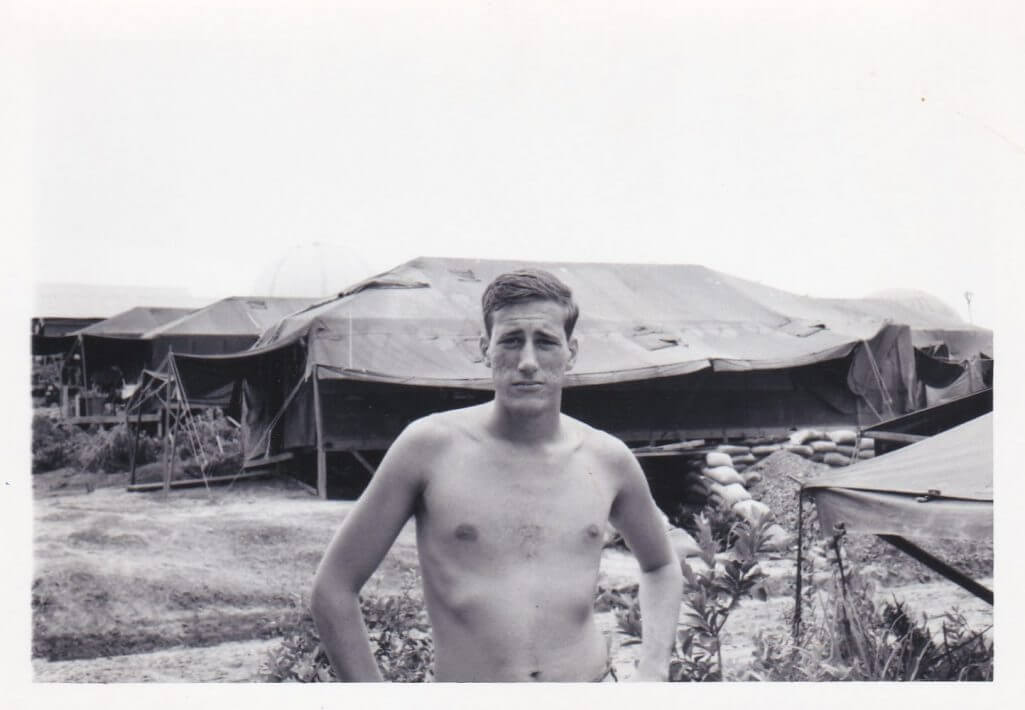 Young soldier standing in front of encampment with shirt off and hands on hips.