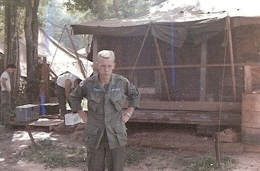 U.S. soldier standing outside an encampment in the jungle with his hands on his hims, looking stern.