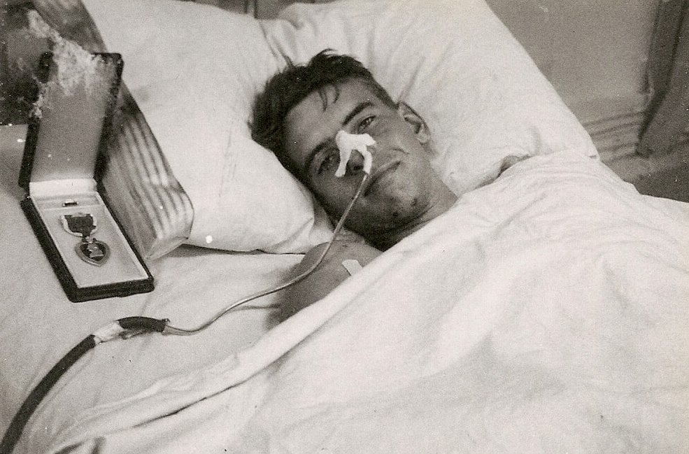 Injured soldier recovering in bed with purple heart next to his pillow.