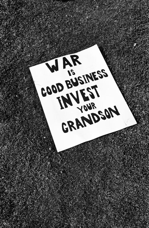 A protest sign lying on the grass: "War is good business. Invest your grandson."