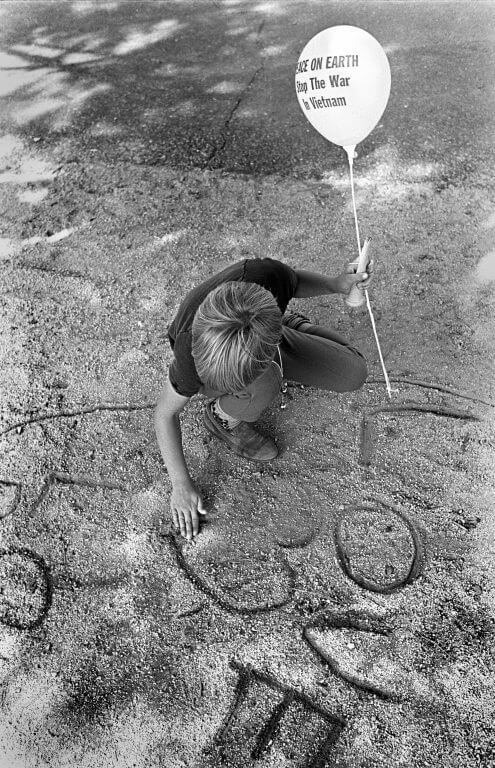 A child writes "Love" in the dirt, holding a white balloon that says "Peace on Earth, End the War in Vietnam."