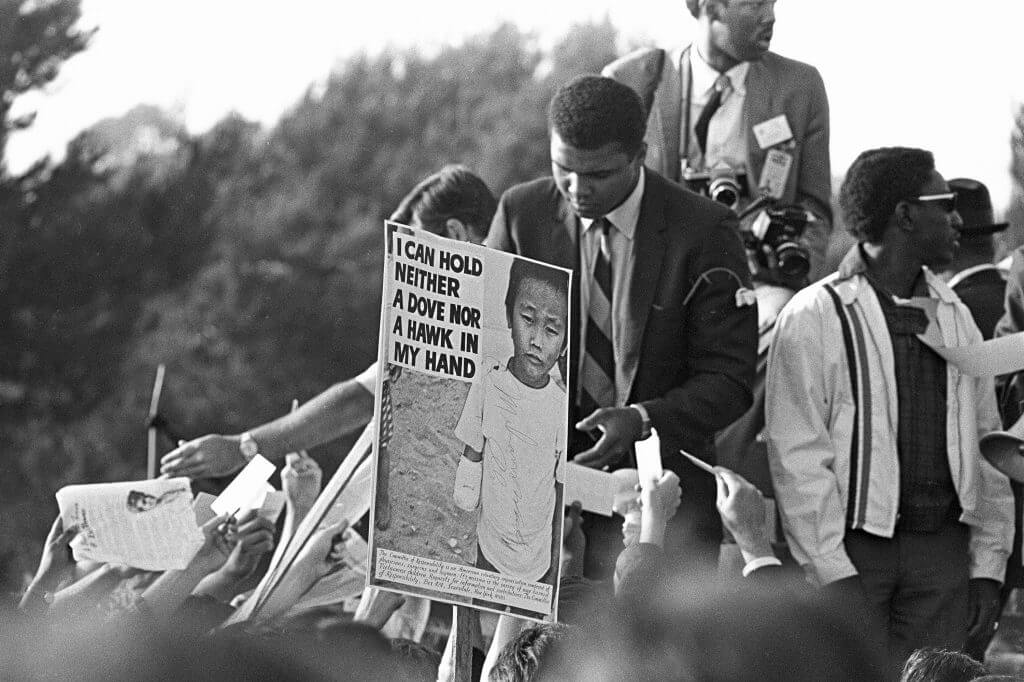 Muhammad Ali signing autographs. Sign in the foreground that features a young Asian child with a bandaged arm: "I can hold neither a dove nor a hawk in my hand."