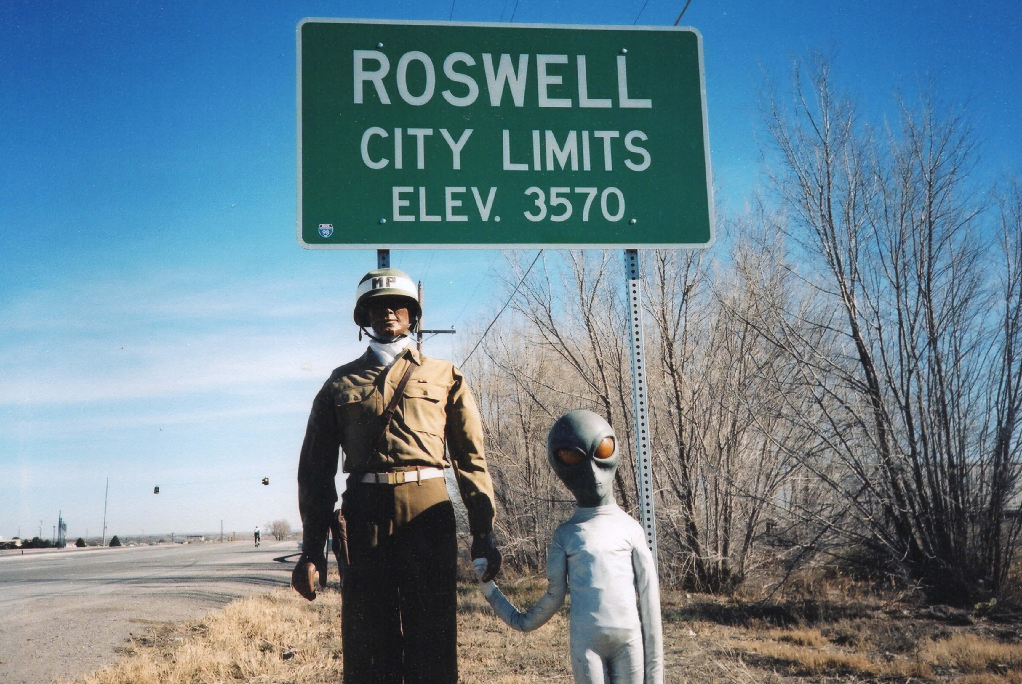 A mannequin in uniform holding the hand of a squat alien figure, in front of a sign that reads "Roswell City Limits, Elev. 3570."