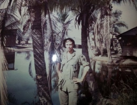 Young soldier standing with hands on hips amid palm trees.