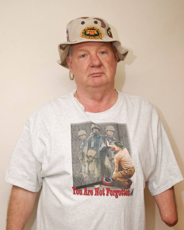 Contemporary photo of an aged U.S. veteran with one arm, looking somber, in a boonie hat with a Vietnam memorial t-shirt.