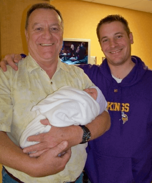A contemporary photo of an older gentleman holding an infant, with a younger man with his arm around him.