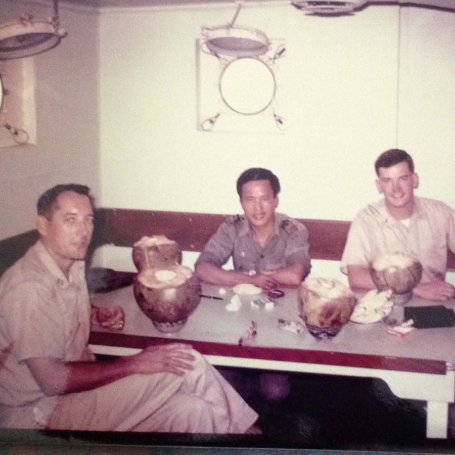 Two U.S. soldiers in khaki uniforms with an Asian man, seated around a table eating coconut.