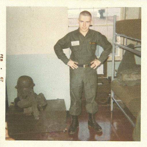 Young soldier with shorn head, standing in barracks with hands on hips.