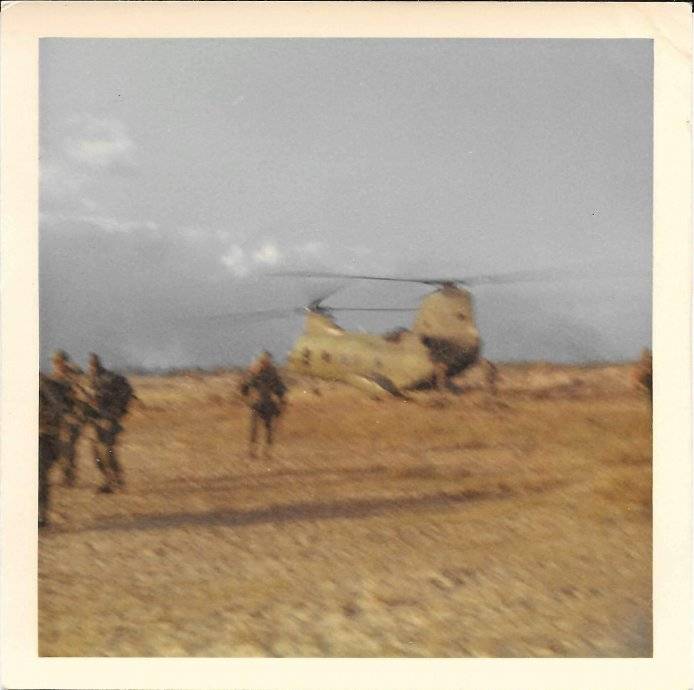 A Chinook helicopter taking off from a field, soldiers stand nearby.