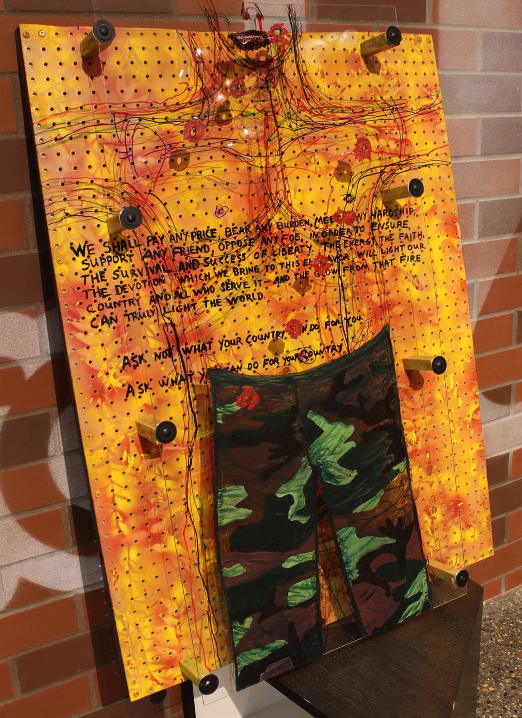 Mixed-media image: pegboard painted orange and red, plexiglass with a shirtless soldier drawn on, bullet holes, and text: We shall pay any price, bear any burden, meet any hardship, support any friend, oppose any foe, in order to ensure the survival and success of liberty..."