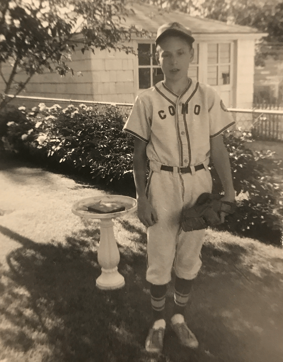 A young boy standing in a yard, wearing his little league uniform. Text across his jersey reads: "COMO."