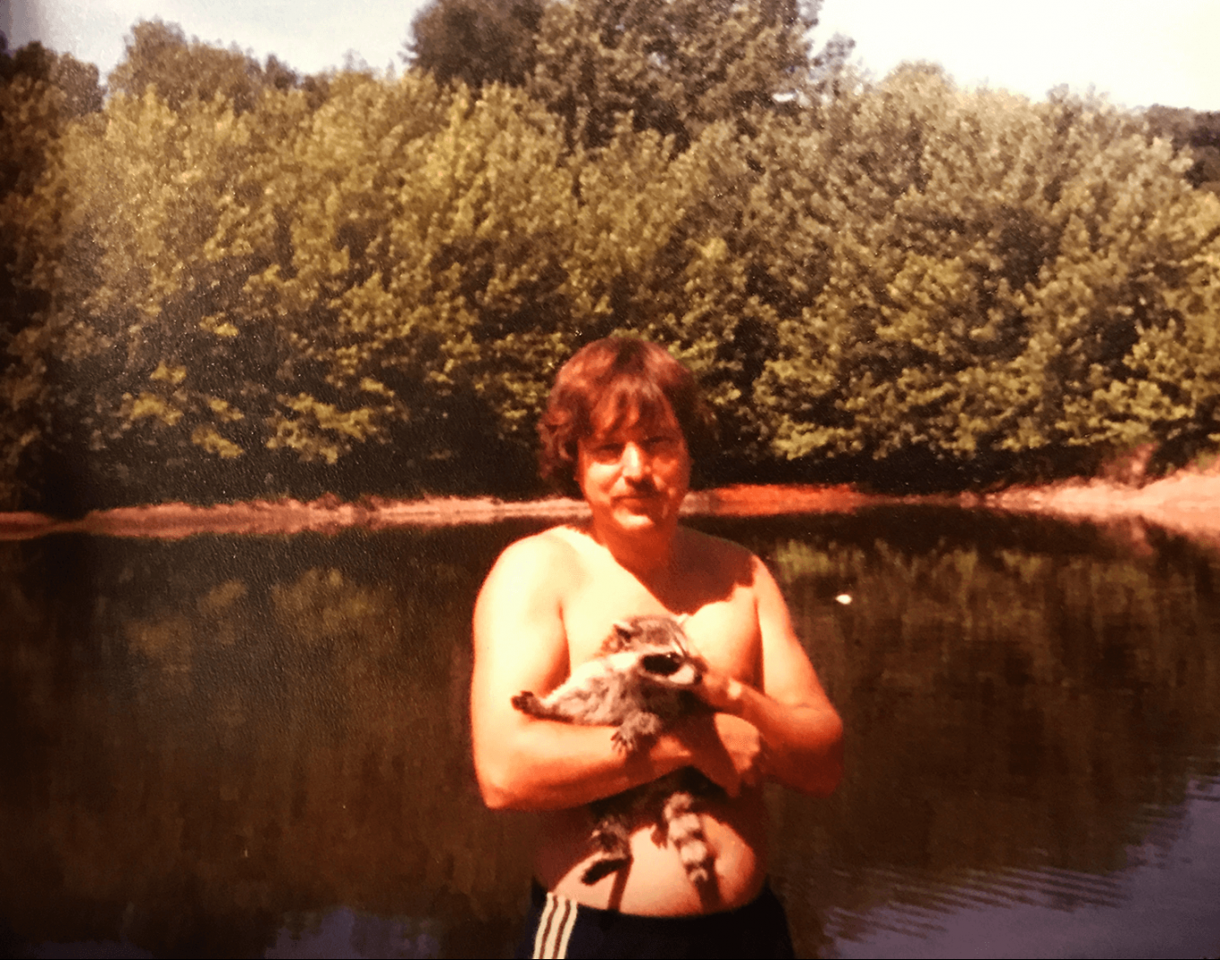 Shirtless man holding a small raccoon, a body of water in the background.