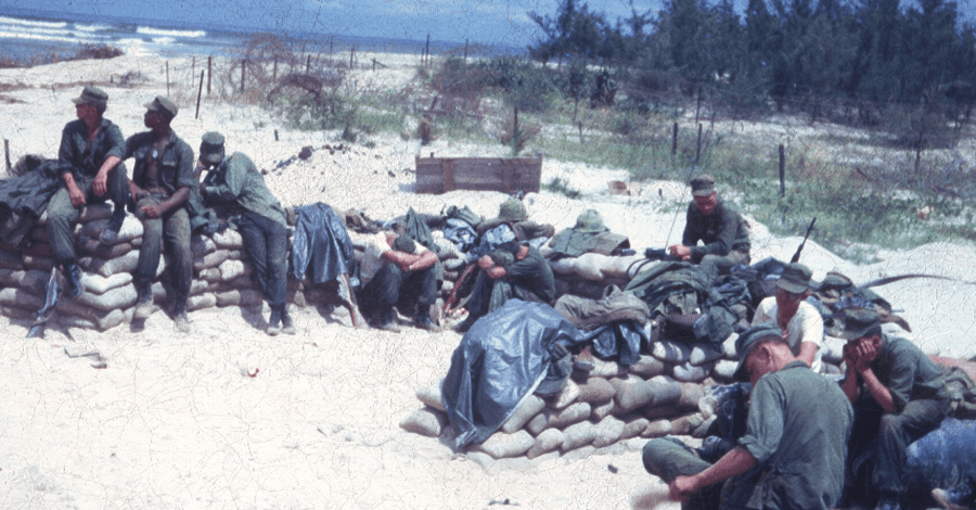 Young soldiers sitting on sandbags on a beach, looking bored.