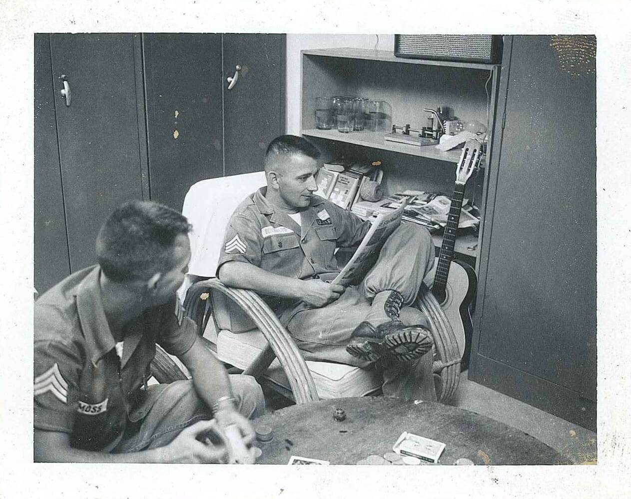 Two U.S. soldiers seated indoors, one reading the paper and the other playing cards.