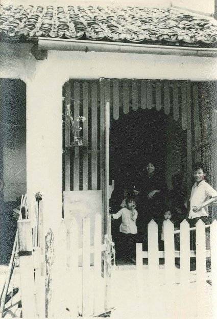 An Asian family standing in a doorway and smiling, white picket fence in the foreground.