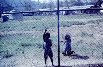 Small Asian children standing on the other side of a chainlink fence.