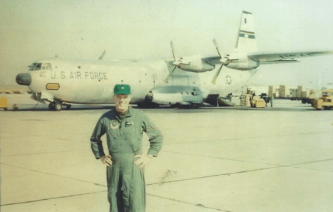 A pilot stands with his hands on his hips on the tarmac in front of a plane.