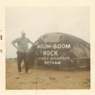 U.S. civilian wearing a construction hat, leaning on a painted rock that reads: "Boom-Boom Rock Monkey Mountain Vietnam." Photo is dated Mar 67.