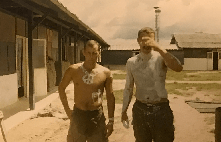 Two shirtless soldiers outside their barracks, with shaving cream on their chests. One is completely covered in cream; one has a peace sign necklace out of cream.