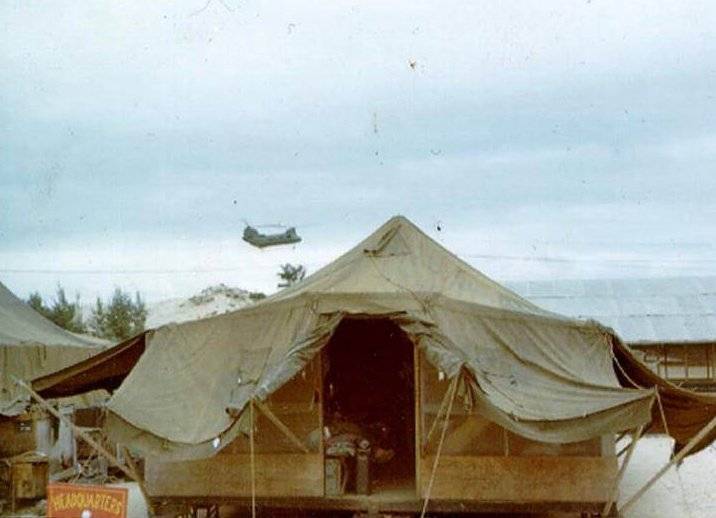 A tent with a "headquarters" sign out front; a Chinook helicopter in the sky behind it.