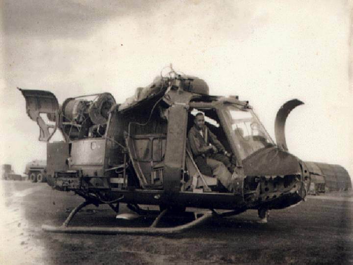 A helicopter without a propeller, on the ground, with a soldier sitting in the co-pilot's seat.