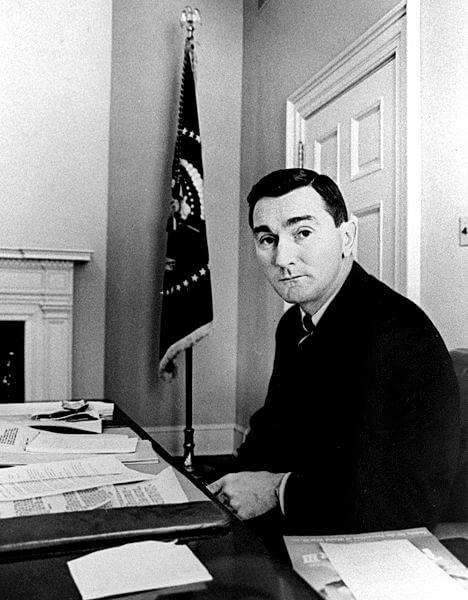 A middle aged man with slicked, black hair sitting at a desk with a flag hanging in the back corner.