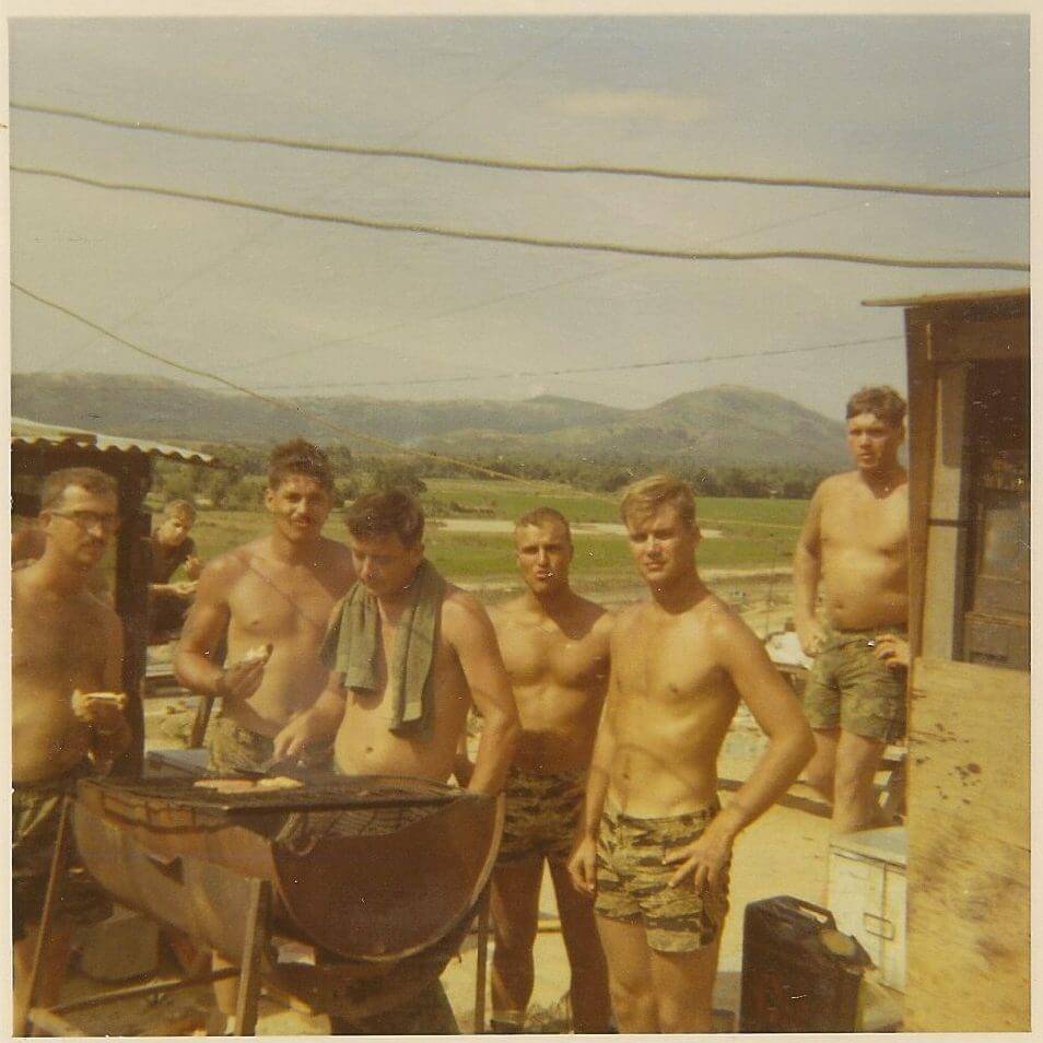 Group of shirtless young U.S. soldiers grilling outside.