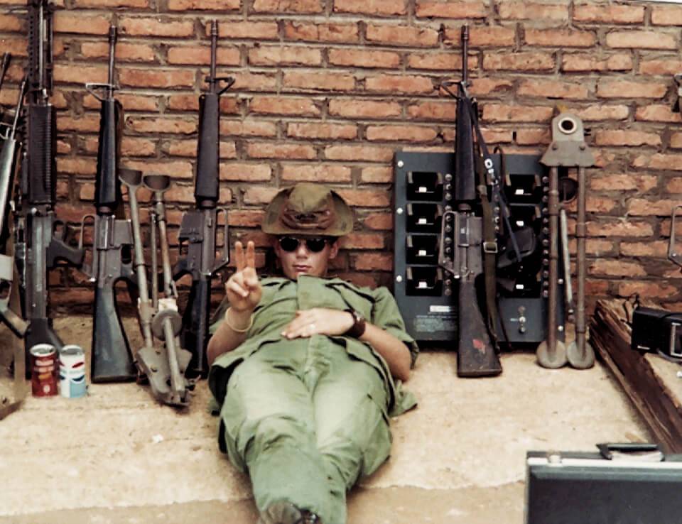 A U.S. soldier slumped against a brick wall with several guns. He wears a bush hat and sunglasses, and holds up a peace sign.
