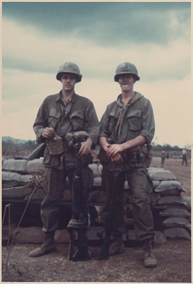 Two young soldiers in full gear, helmets; standing in front of a row of sandbags.