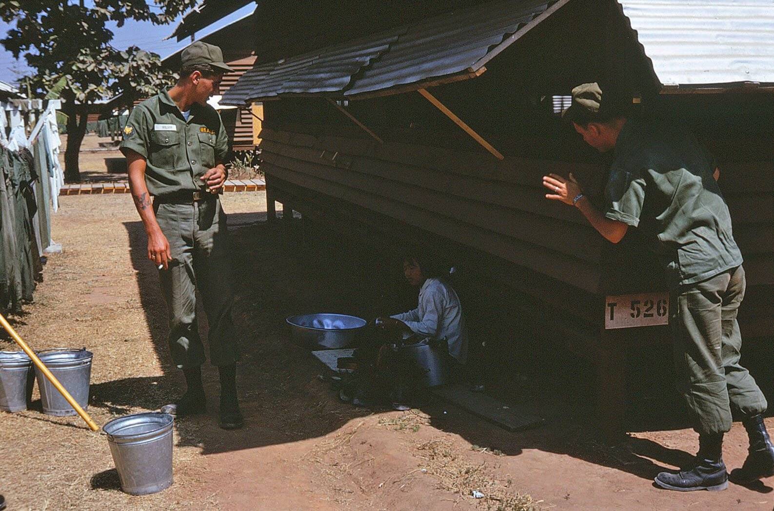 Two US soldiers outside, talking to an Asian woman sitting in the shade, hand washing her laundry.