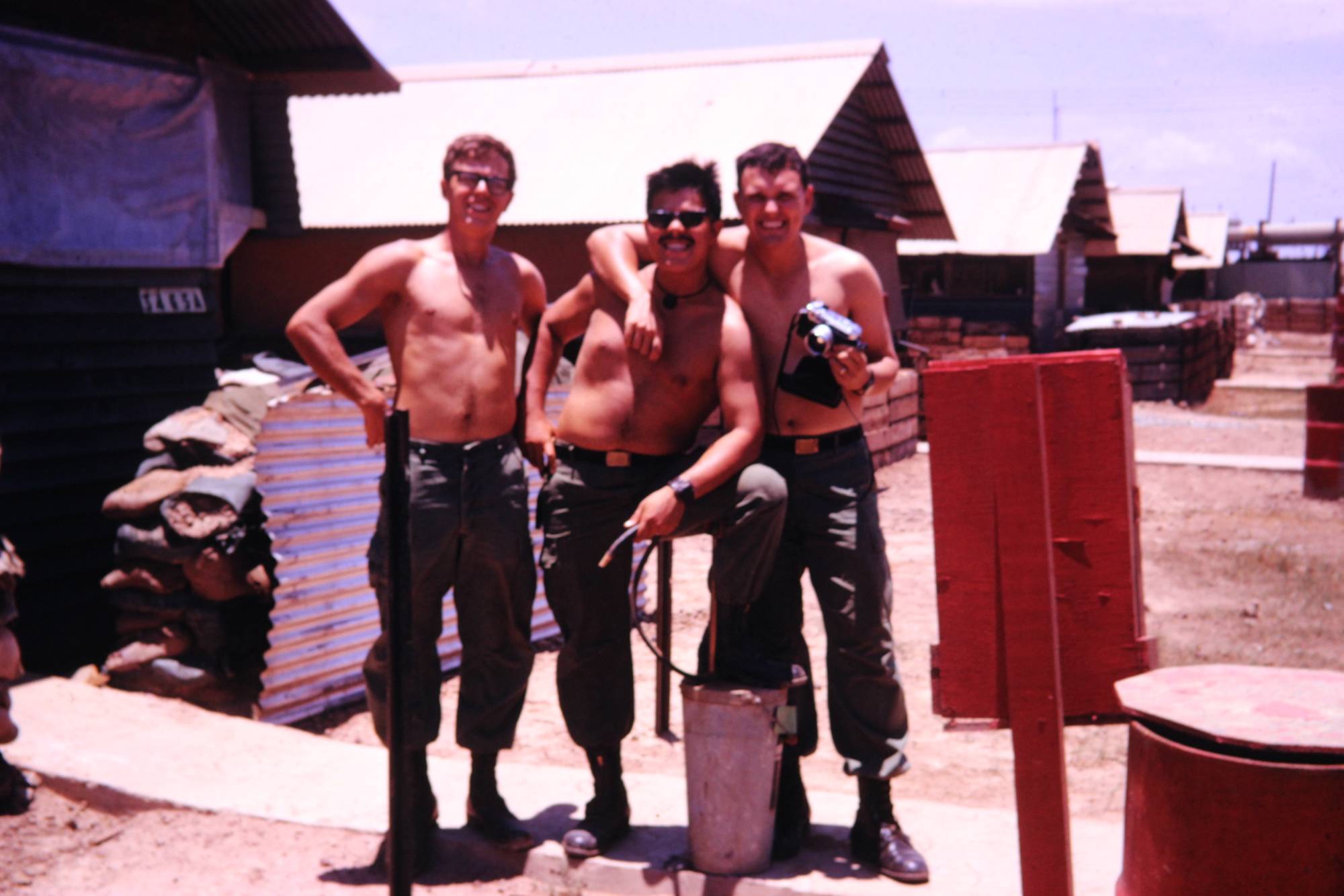 Three shirtless soldiers outside temporary living quarters.